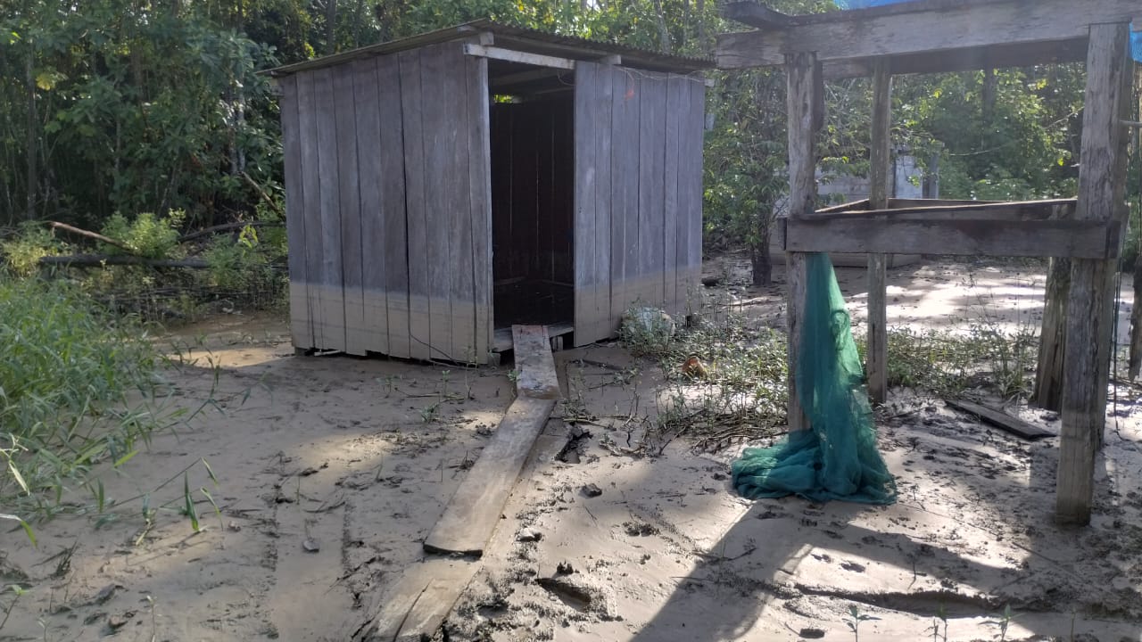 Um banheiro de madeira e coberto de telha a frente uma ponte improvisada de madeira, marca da água aproximadamente meio metro, cercado por lama em uma área ribeirinha, cercada por vegetação.