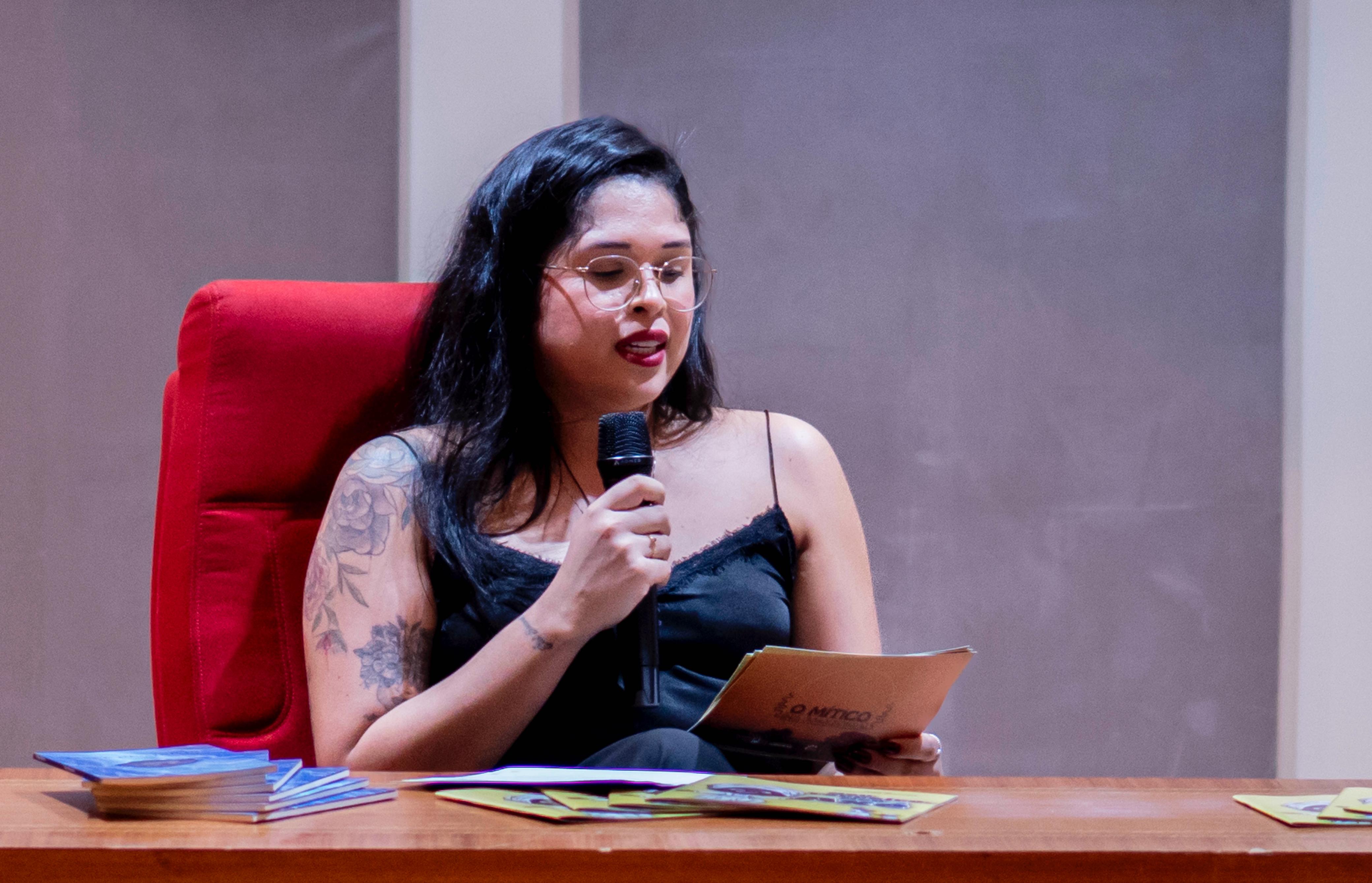 Fotografia colorida. Monique Pereira mulher está sentada em uma poltrona vermelha, segura um microfone com a mão direita e um papel na mão esquerda, enquanto lê algo para o público. Ela está vestida com um top preto de alças finas, usa óculos de armação clara e possui uma tatuagem floral no braço esquerdo. À sua frente, sobre a mesa de madeira, estão dispostos materiais impressos.
