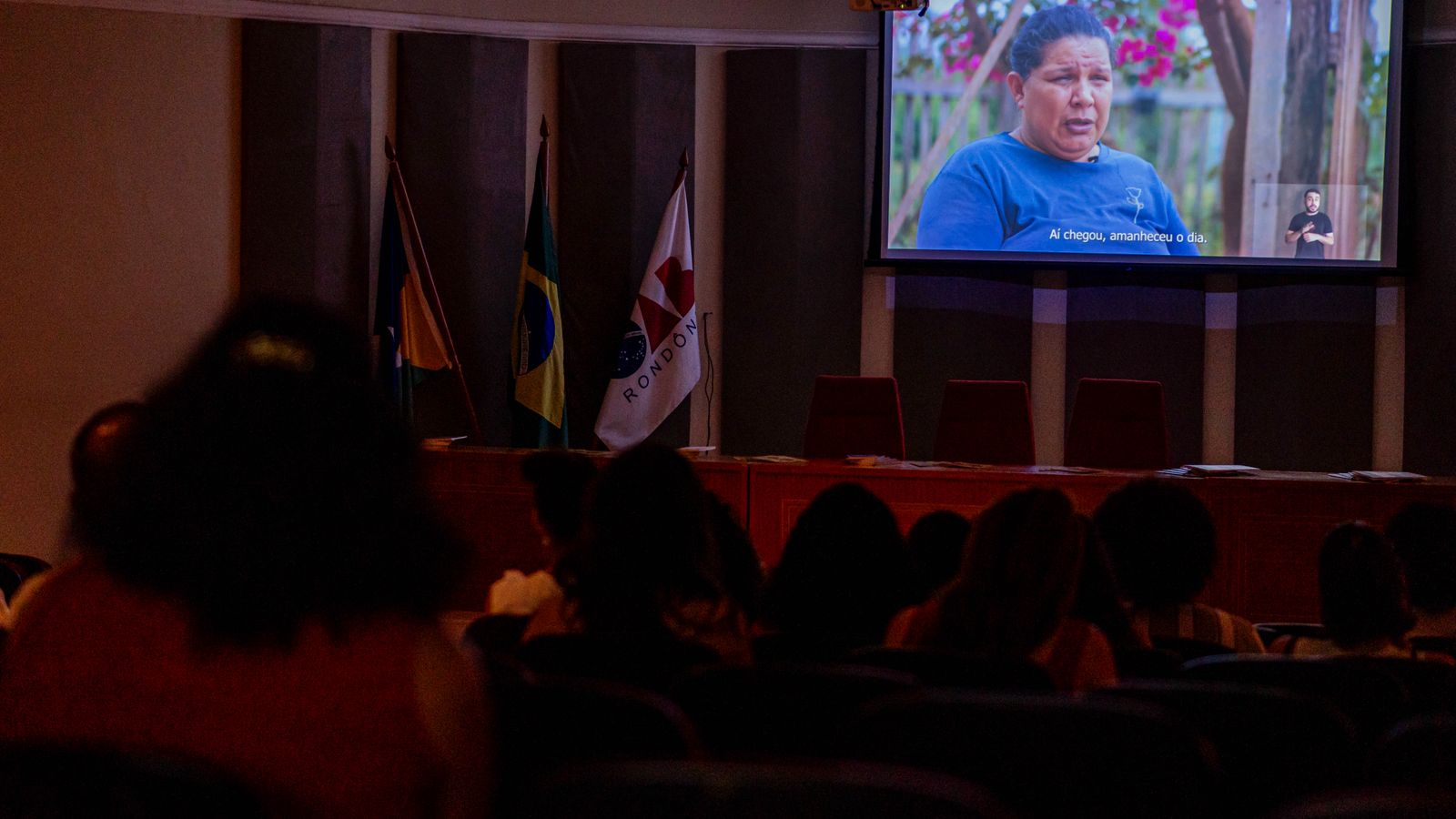 Fotografia colorida. Auditório da Ordem dos Advogados do Brasil na sede de Rondônia durante a exibição do documentário. No telão, uma mulher compartilha sua história, enquanto o público acompanha atento. Ao lado esquerdo do telão estão as bandeiras do Brasil, de Rondônia e da Ordem dos Advogados de Brasil. 