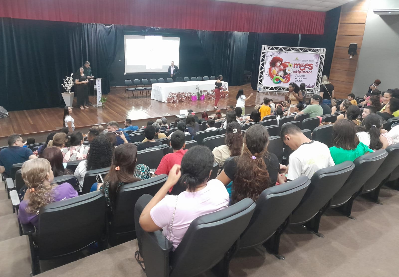 Fotografia colorida de um auditório com cerca de 100 pessoas sentadas em poltronas de couro preto, voltadas para o palco. No palco, 4 pessoas em pé, em um ambiente com paredes e piso escuros e cortinas pretas com bordas vermelhas. O palco apresenta um painel com a mensagem 'Mães atípicas: agora é com você'. Ao centro, uma mesa coberta com uma toalha branca,3 jarro de flores e aproximadamente 15 presentes à esquerda bicicleta embrulhadas. 