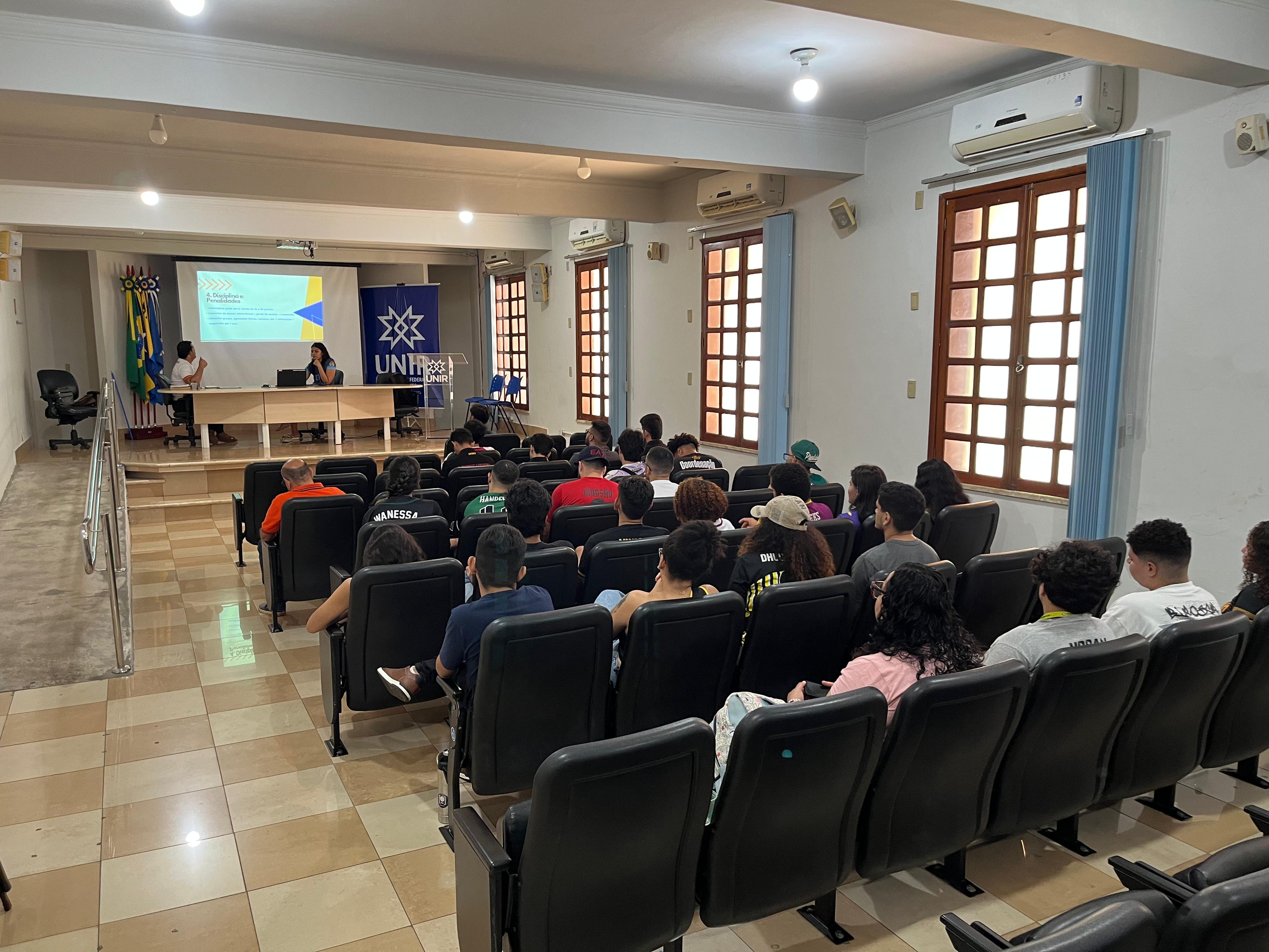 alunos sentados em um auditório, à frente uma mesa com 2 instrutores sentados, apresentando um slide.
