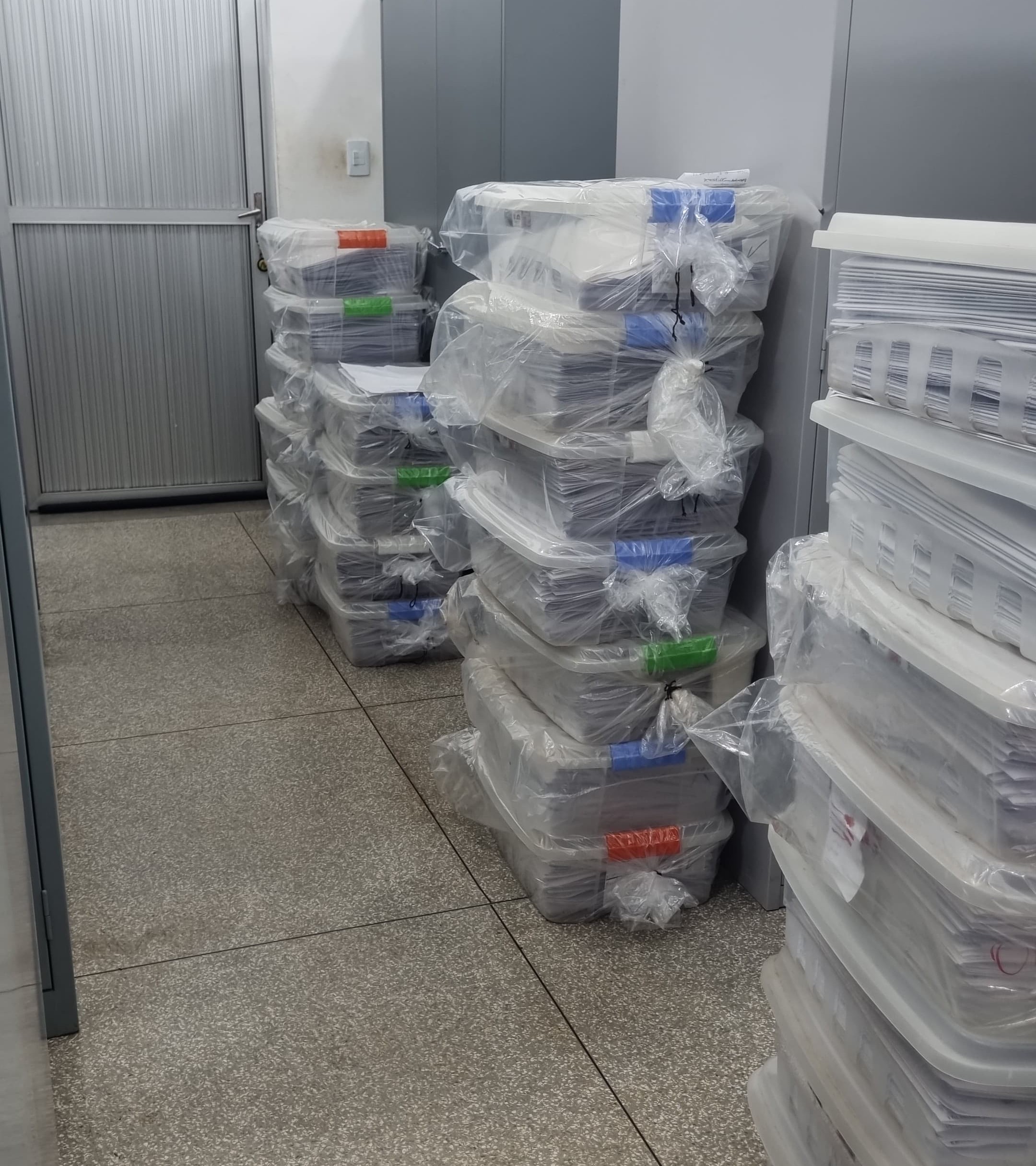 A imagem mostra um laboratório de arquivamento de biologia. No ambiente, há armários metálicos cinza e diversas caixas plásticas empilhadas, todas envolvidas em sacos plásticos transparentes. Dentro das caixas, há arquivos organizados em grande volume. O chão é de granilite e, ao fundo, há uma porta metálica fechada.