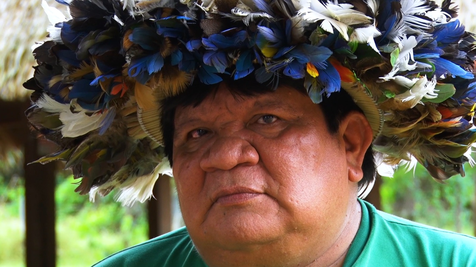 Almir Suruí, liderança do povo Paiter Suruí, usando um cocar feito com penas coloridas de diversas aves. Ele veste uma camisa verde e olha com expressão séria para a câmera. Ao fundo, vê-se uma estrutura de madeira e vegetação da floresta amazônica.