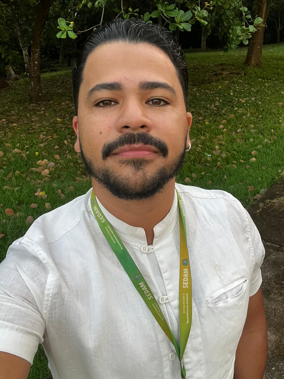 Na imagem, um homem aparece em um ambiente externo, com grama e árvores ao fundo. Ele está usando uma camisa branca de linho com gola padre e botões frontais, além de um crachá verde e amarelo com a inscrição “SEDAM”. Ele olha diretamente para a câmera com expressão serena.