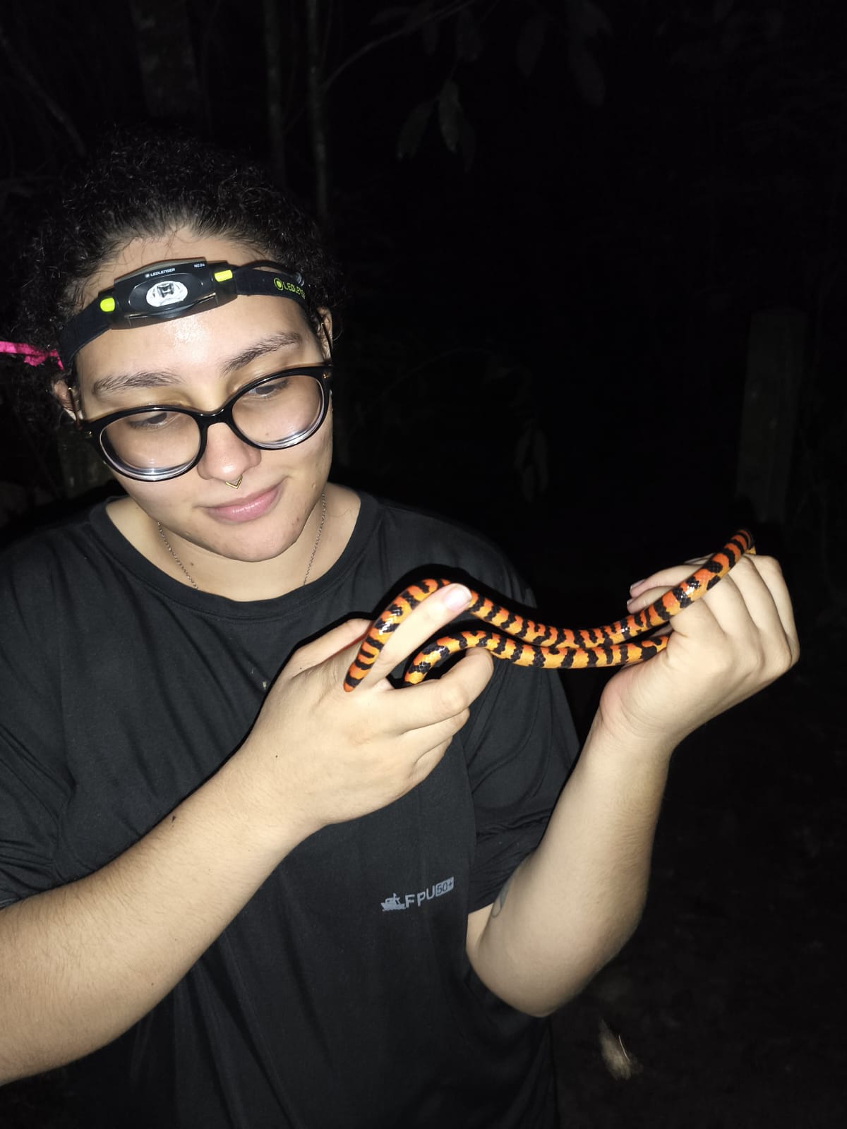 Foto noturna de uma jovem segurando uma serpente de coloração vibrante, com anéis laranjas, pretos e amarelos. Ela usa óculos de grau, tem cabelopreso e está com uma lanterna de cabeça. Veste uma camiseta preta. A iluminação foca nela, enquanto o fundo está escuro, sugerindo floresta ao redor.