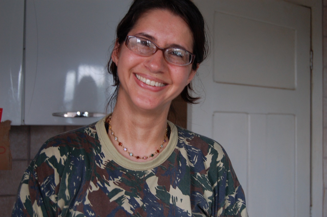 Foto de uma mulher sorridente em um ambiente interno. Ela usa óculos de grau, colar com pequenas pedras coloridas e camiseta camuflada em tons de verde e marrom. Seus cabelos escuros estão presos para trás. Ao fundo, há uma porta branca e parte de um armário de cozinha. A iluminação indica que é dia.