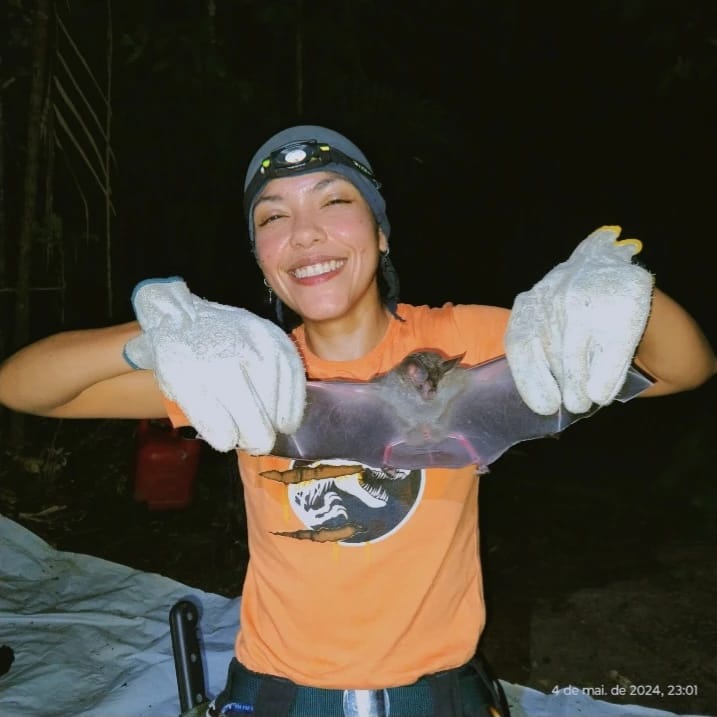 Foto noturna de uma jovem sorridente, segurando cuidadosamente um morcego com as asas abertas. Ela usa luvas grossas, camiseta laranja com estampa de animal, calça escura, touca e lanterna de cabeça. A cena ocorre em área de floresta, com lona branca no chão ao fundo, iluminada por lanterna.