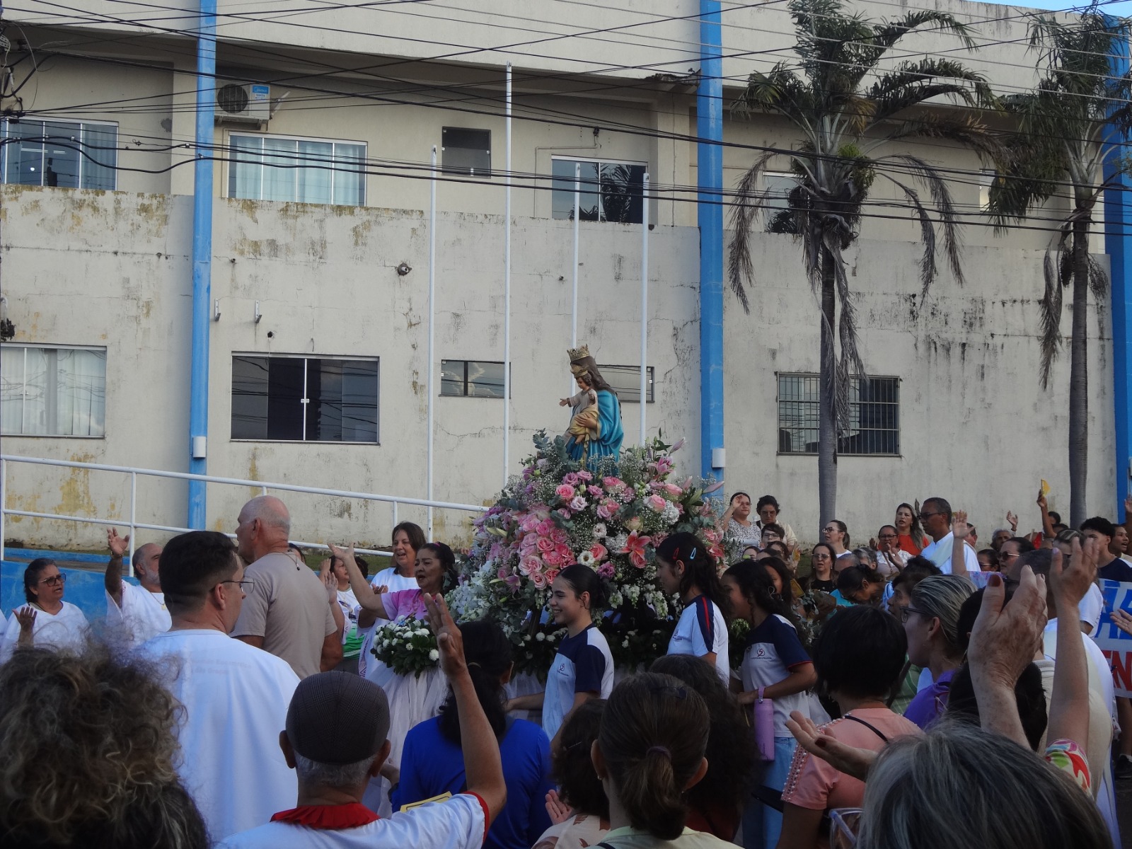 Fotografia colorida. Em primeiro plano e no centro da imagem, uma estátua de Maria Auxiliadora posicionada em um altar de flores. Homens e mulheres rezam com as mãos levantadas. Alguns de olhos fechados, outros de olhos abertos.