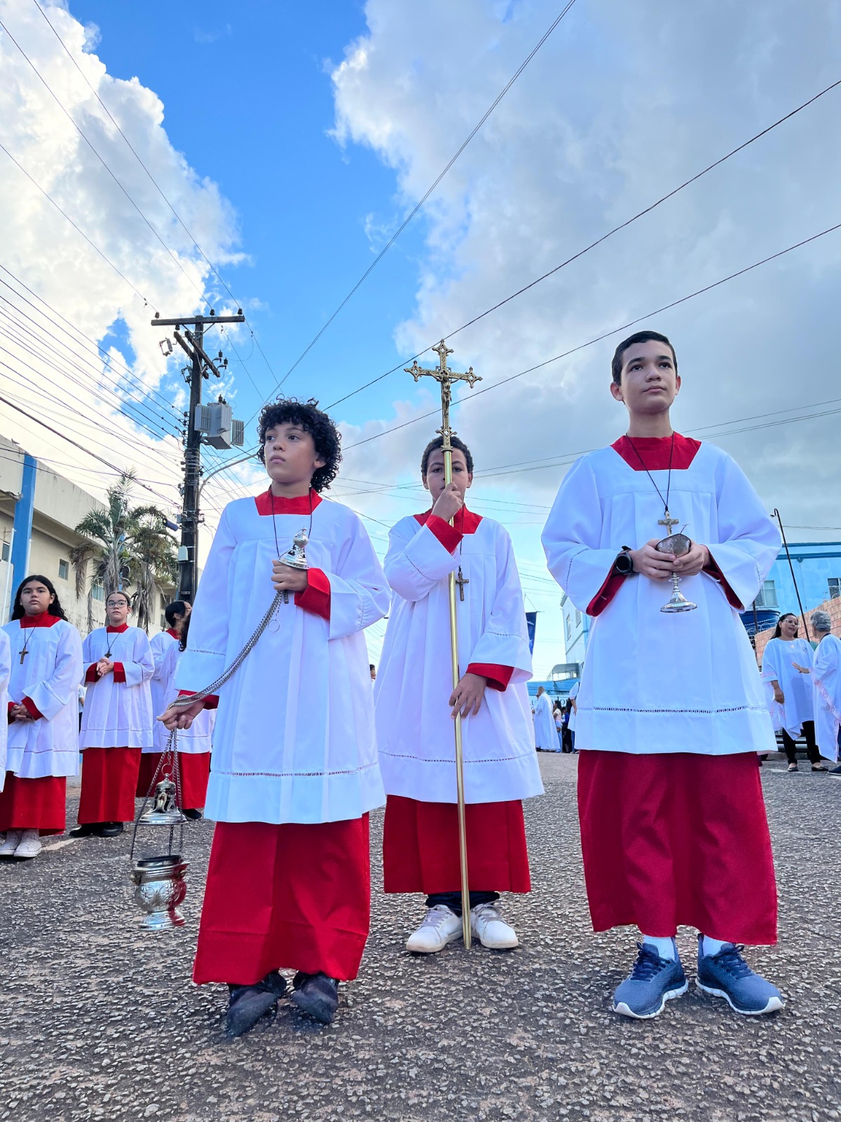 Fotografia colorida. Da esquerda para a direita. Três garotos trajando batinas católicas vermelho e branco. Eles carregam um incensário, uma cruz e um cálice dourados. No segundo plano, céu azul com algumas nuvens.
