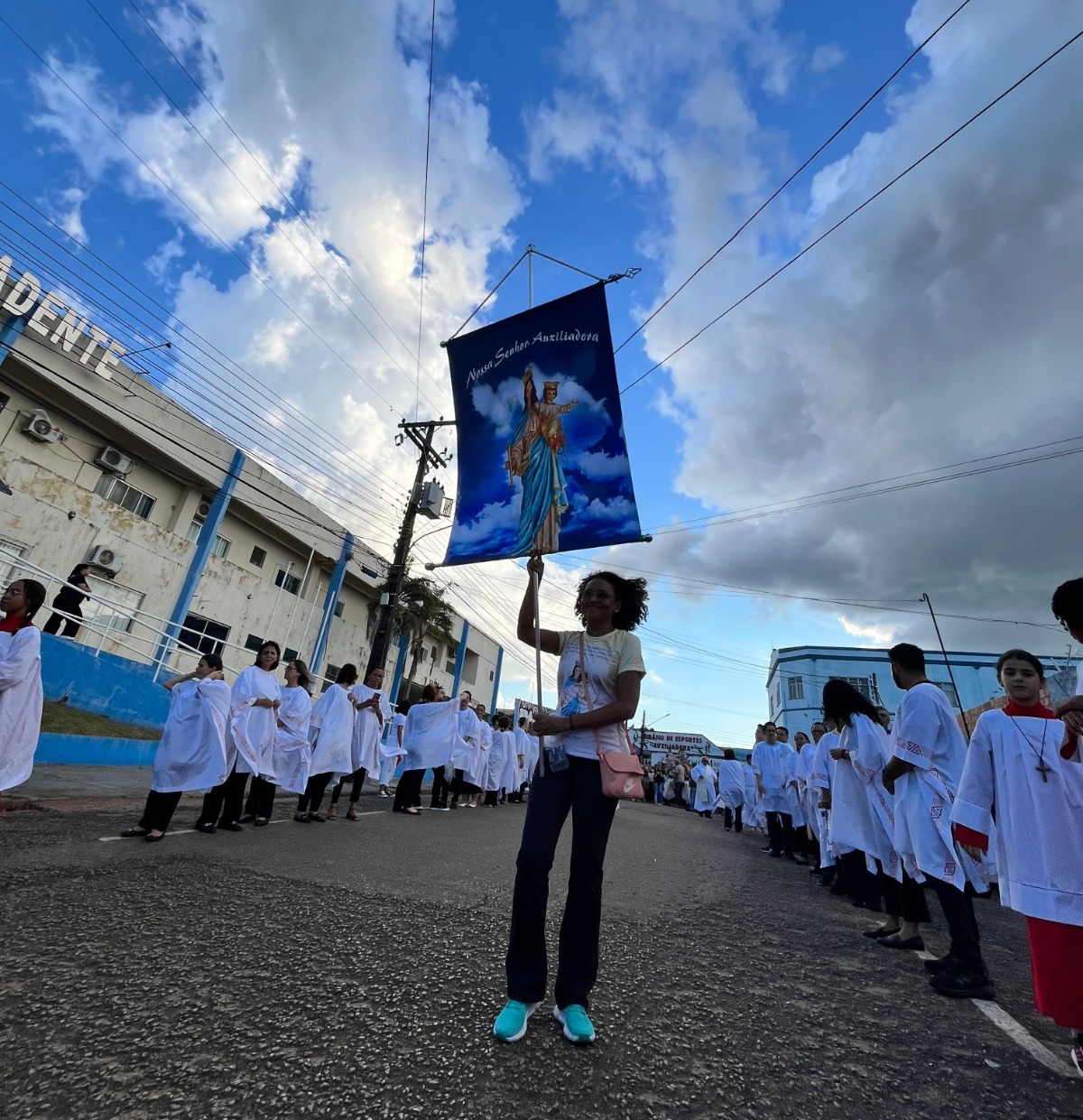 Fotografia colorida. Ângulo alto em contraluz. No centro do primeiro plano, uma mulher com um estandarte de uma santa. Ao lado, coroinhas enfileirados. Céu azul no plano de fundo.