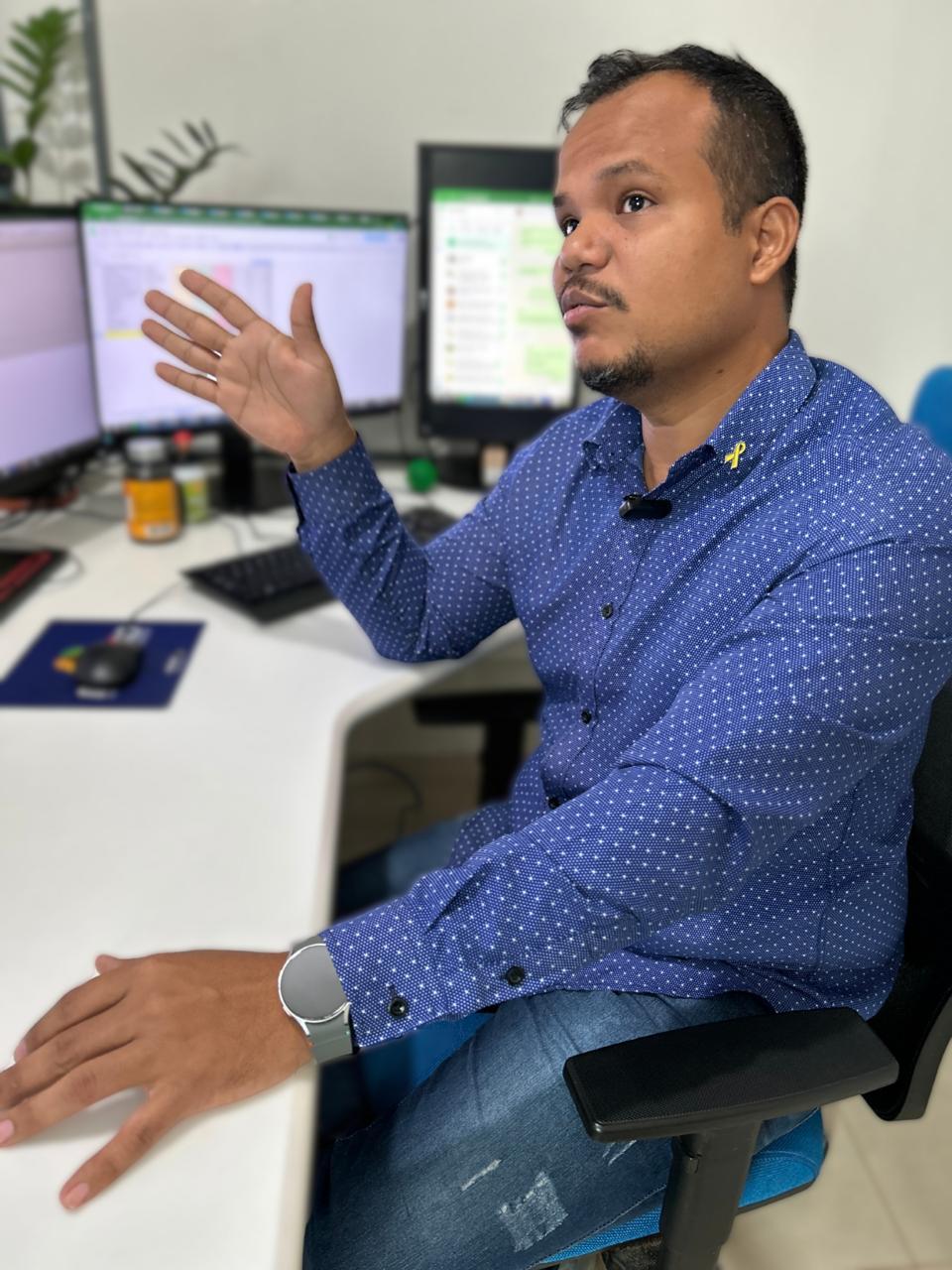 Fotografia colorida. Homem pardo sentado em frente a uma mesa de escritório, usando camisa azul com pontilhado branco e calça jeans. Ele gesticula com a mão direita enquanto conversa, olhando para o lado. Sobre a mesa há dois monitores de computador ligados, teclado, mousepad com mouse, frascos pequenos e outros itens de trabalho. O ambiente é bem iluminado.