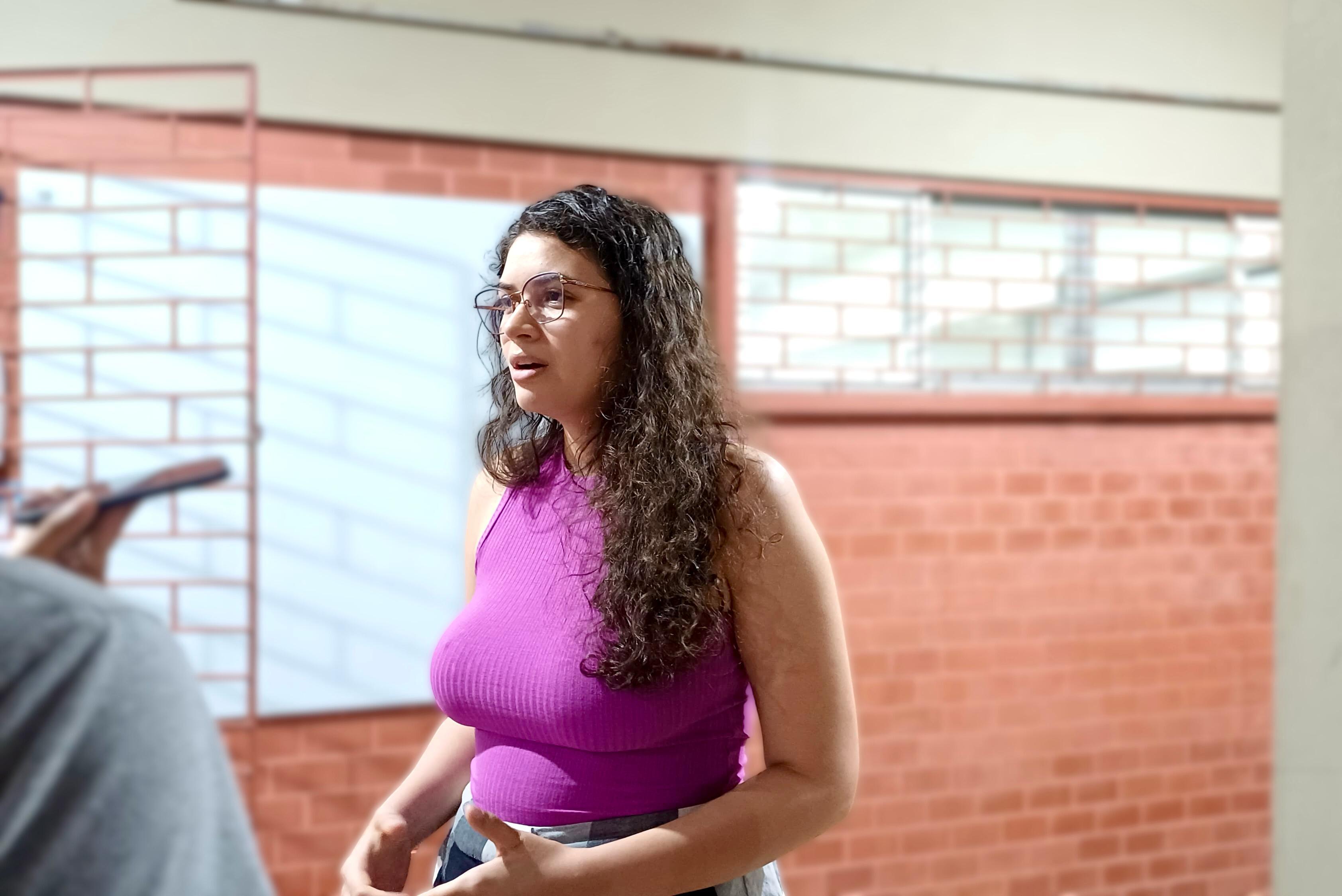 Mulher de cabelos cacheados e óculos fala com expressividade enquanto é entrevistada em frentea uma parede de tijolos. Ela veste uma blusa sem mangas de cor roxa. A mulher demonstra envolvimento e emoção com a conversa. 