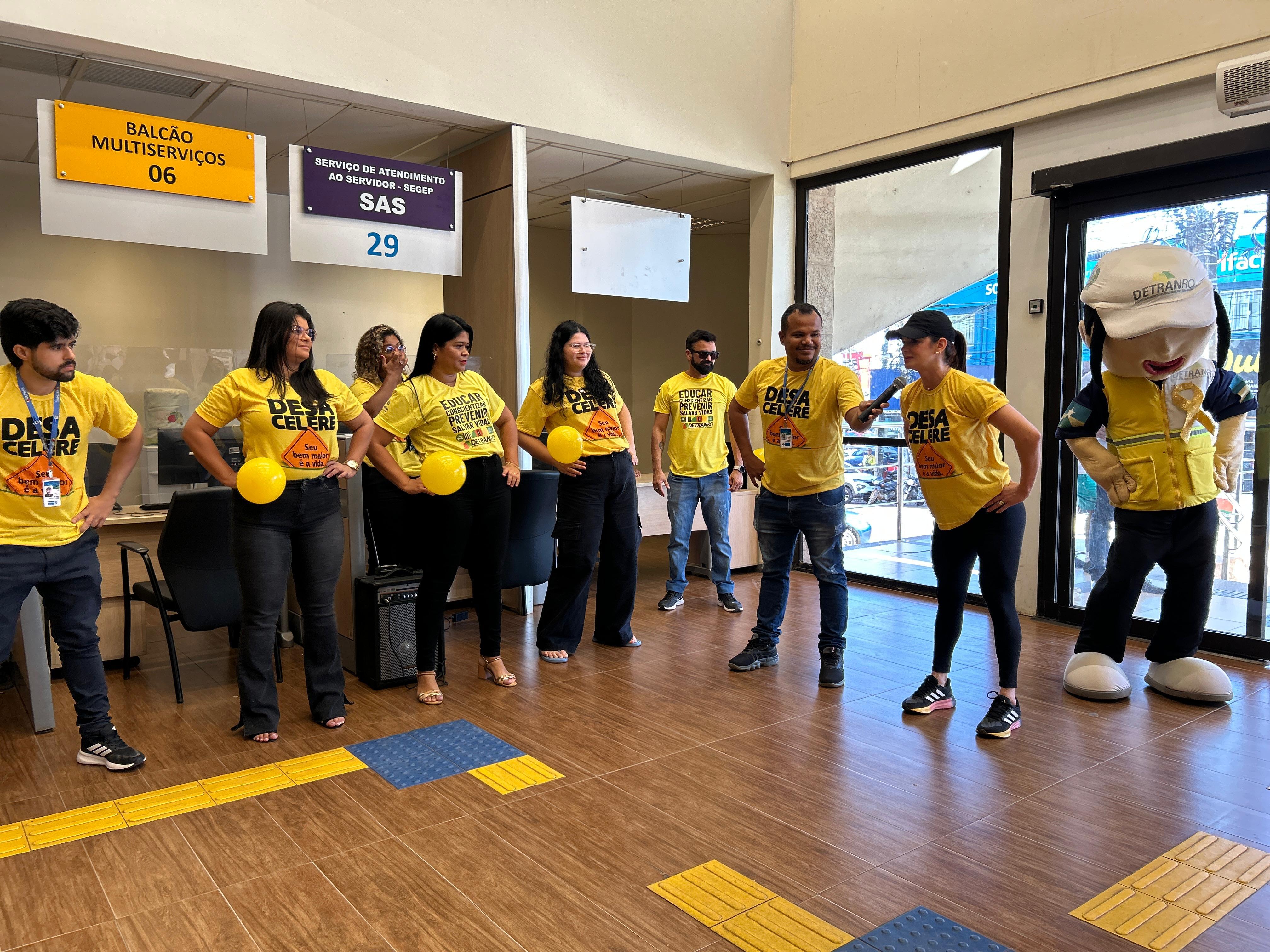 Fotografia colorida. Um grupo de pessoas está reunido em um ambiente interno. Todos usam camisetas amarelas e seguram balões amarelos, demonstrando um clima descontraído e de confraternização. Ao fundo, há uma grande janela que deixa entrar luz natural, e à direita uma pessoa está fantasiada como o mascote “a Vidinha”, o que traz um tom lúdico ao encontro. A atmosfera refere-se a uma ação educativa/campanha, voltada para a segurança no trânsito