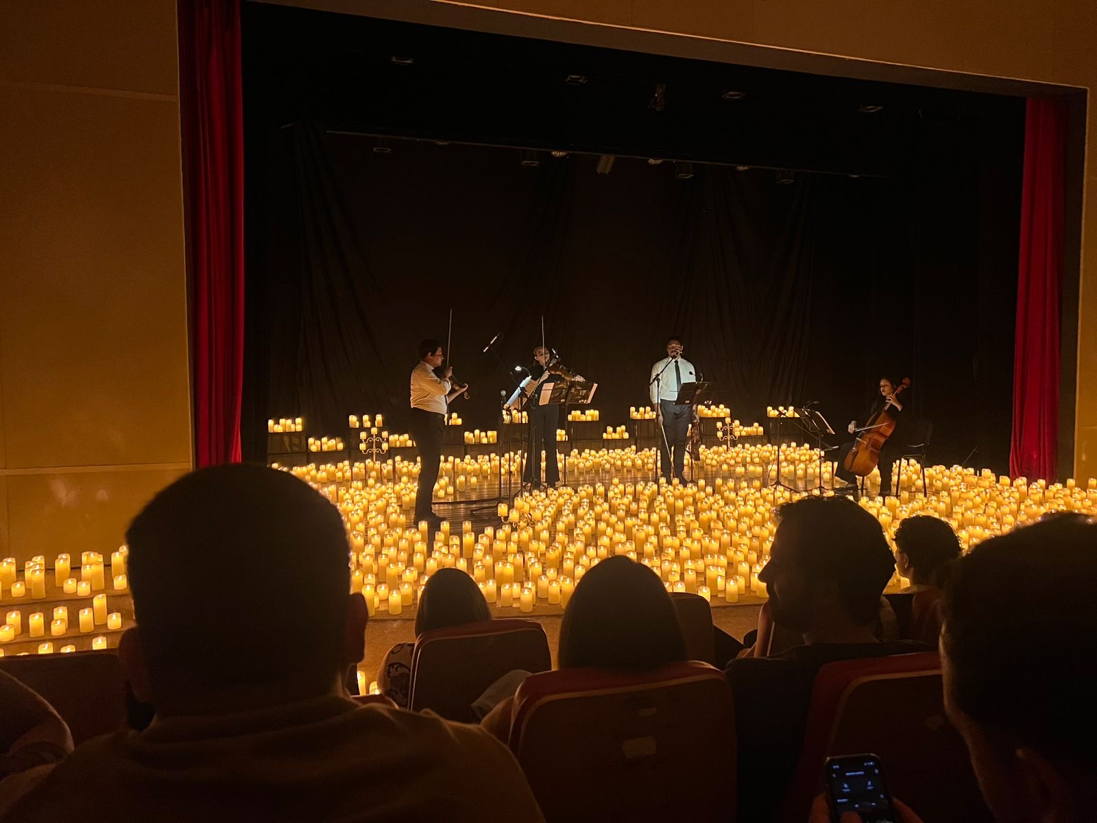 Fotografia colorida de um concerto Candlelight realizado no Teatro Guaporé, em Porto Velho. Quatro músicos tocam instrumentos de corda: dois violinos, uma viola de arco e um violoncelo. Todos estão sobre um palco coberto por centenas de velas eletrônicas acesas, criando uma atmosfera acolhedora e intimista. A plateia assiste em silêncio, com a iluminação do ambiente focada nas velas e nos artistas.