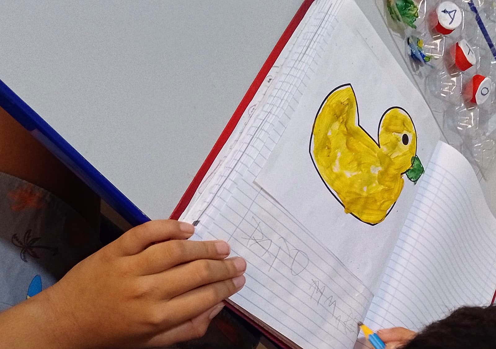 Fotografia colorida. Uma criança segura um caderno aberto sobre a mesa. Na página, há um desenho de um pato pintado de amarelo, com contorno preto e bico verde. Abaixo da imagem, a criança escreveu com lápis: "PATO AMARELO". Ao lado do caderno, há tampinhas com letras feitas de papel e plástico. A mão da criança aparece segurando um lápis azul.