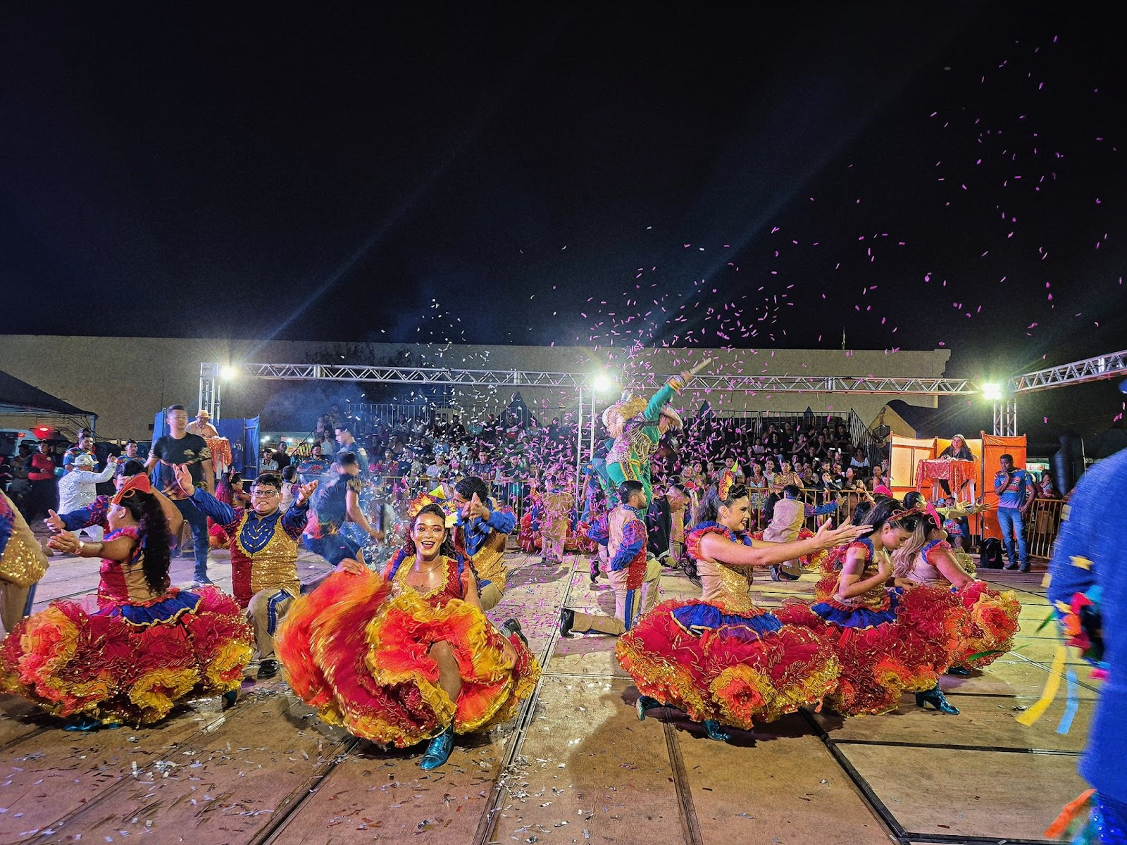 A fotografia colorida mostra uma apresentação típica de festa junina, com um grupo de dançarinos vestidos com trajes coloridos e tradicionais de quadrilha. Eles estão em um palco ao ar livre, cercados por luzes e fumaça cenográfica, enquanto confetes caem do alto, criando um clima festivo. O público pode ser visto ao fundo assistindo ao espetáculo.